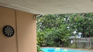 An outdoor patio with a wicker table, a sofa with yellow cushions, a dartboard, and a pool in the background. - Cozy 2-bedroom villa with Pool in sunny Miami (Miami)