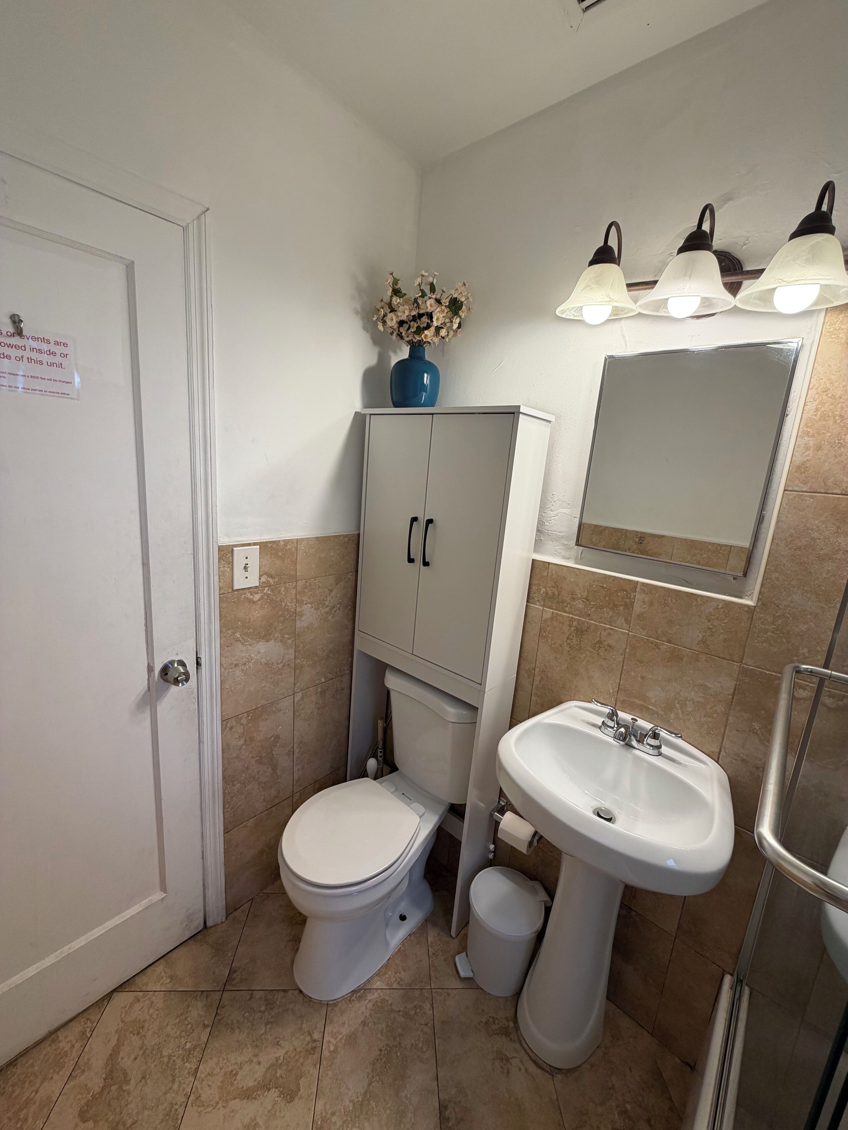 A bathroom with a toilet, a pedestal sink, a cabinet, a mirror, and a vase with flowers on the wall.
