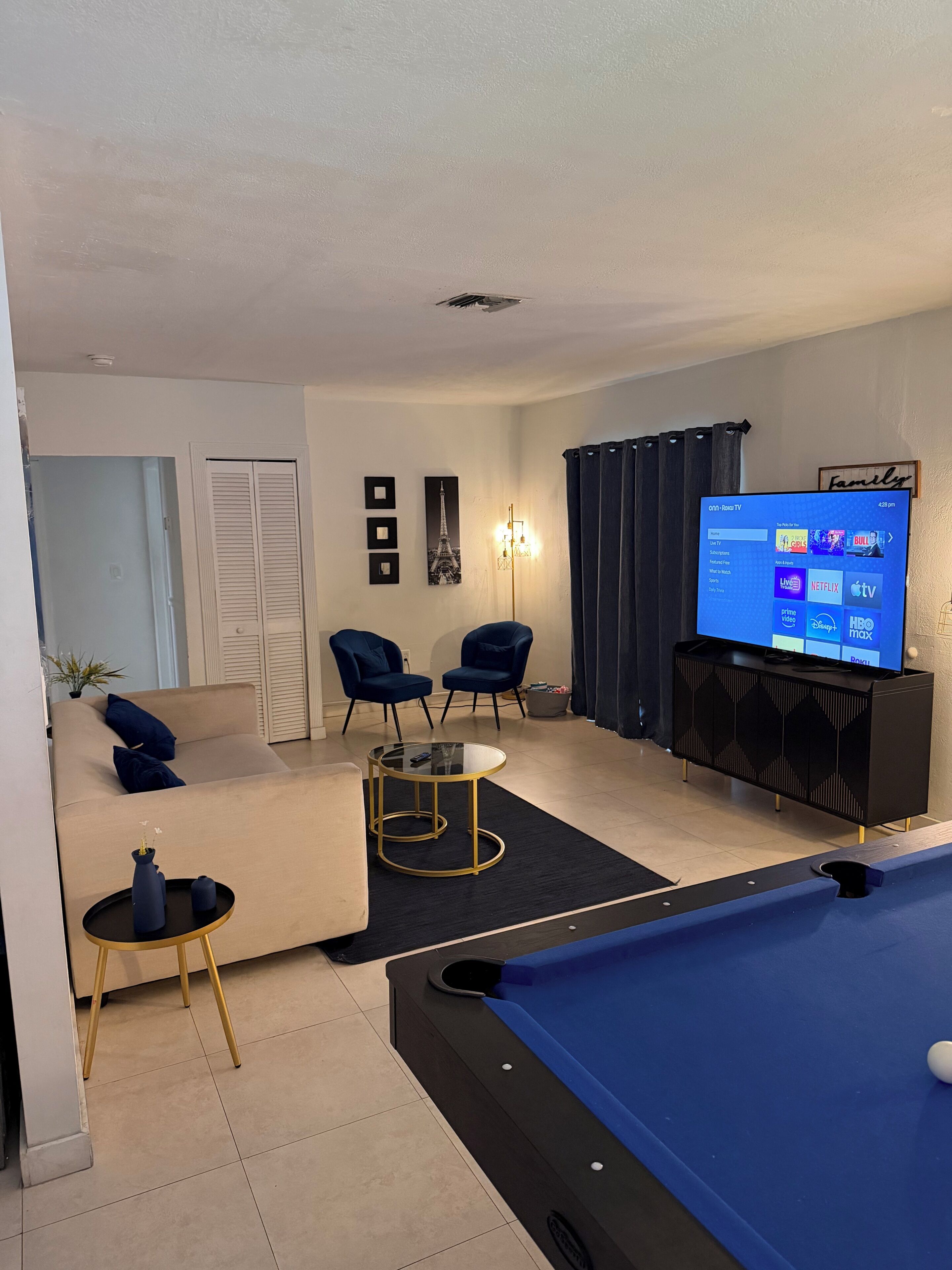 Ein Wohnzimmer mit Billardtisch, einer Couch, zwei Sesseln, einem Fernseher, der einen Streaming-Dienst zeigt, und einem gerahmten Bild vom Eiffelturm.
