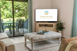 Ein Wohnzimmer mit einem Fernseher, der 'GRAND WELCOME BRANSON & THE OZARKS' zeigt, einem Glastisch mit Stühlen, einer gepolsterten Bank und einem Holzschrank mit einer Pflanze.