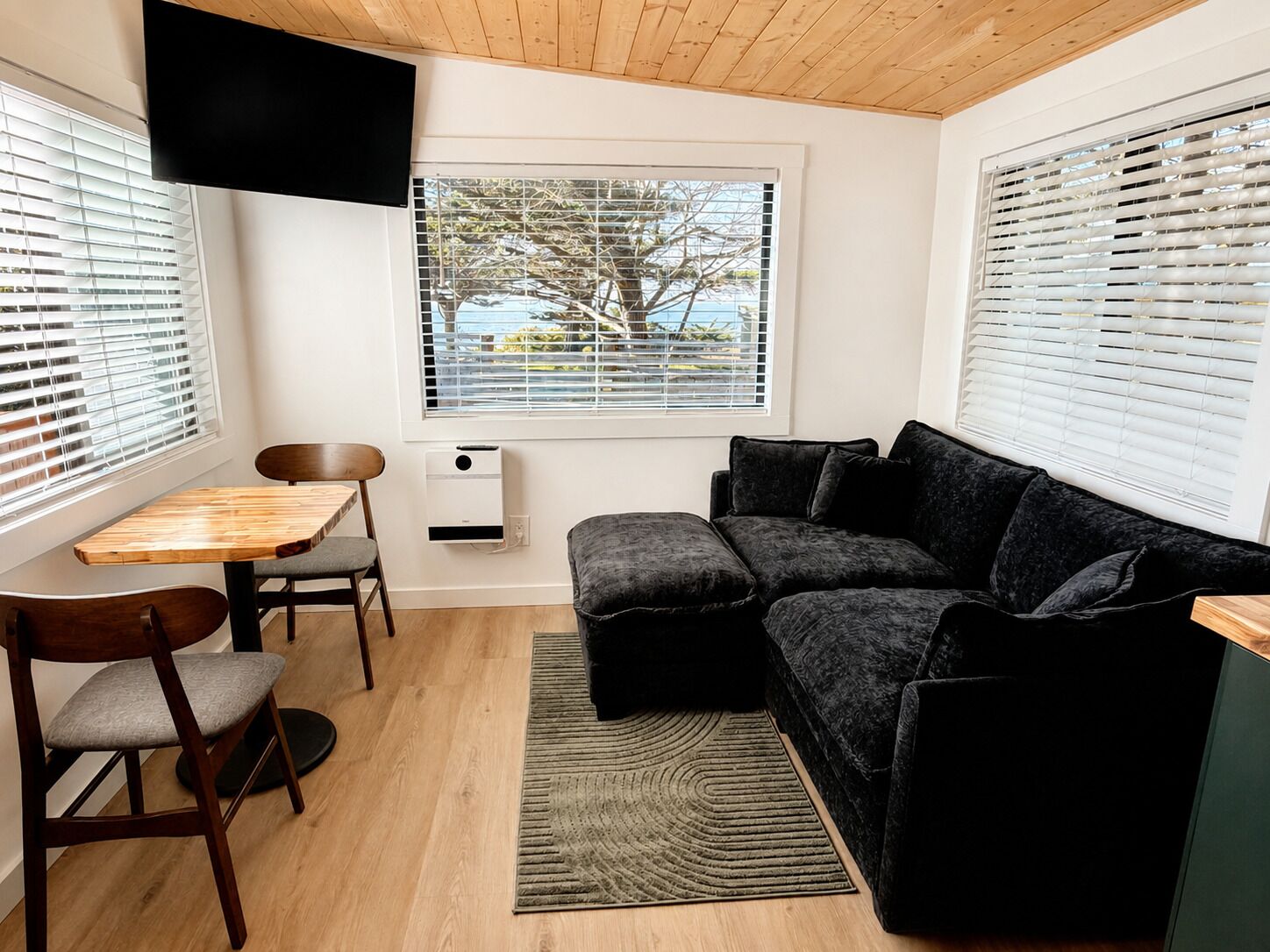 Un espacio habitable compacto con techo de madera, un sofá negro, una pequeña mesa con sillas y una ventana con persianas.