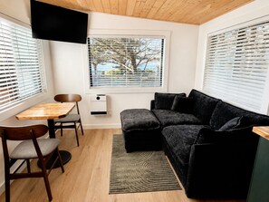 Un espace de vie compact doté d’un plafond en bois, d’un canapé noir, d’une petite table avec des chaises et d’une fenêtre avec des stores.