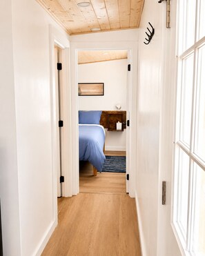 Un pasillo estrecho que conduce a un dormitorio con cama, mesita de noche y estante en la pared.