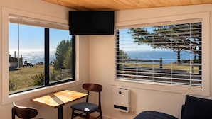 A room with a wooden ceiling, a TV mounted on the wall, a table with two chairs, and a view of the ocean through large windows.