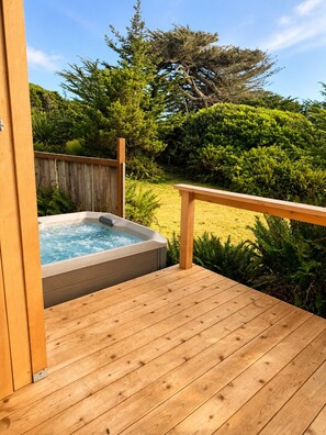 Une terrasse en bois avec un jacuzzi et une vue sur un jardin où poussent différentes plantes et un arbre.