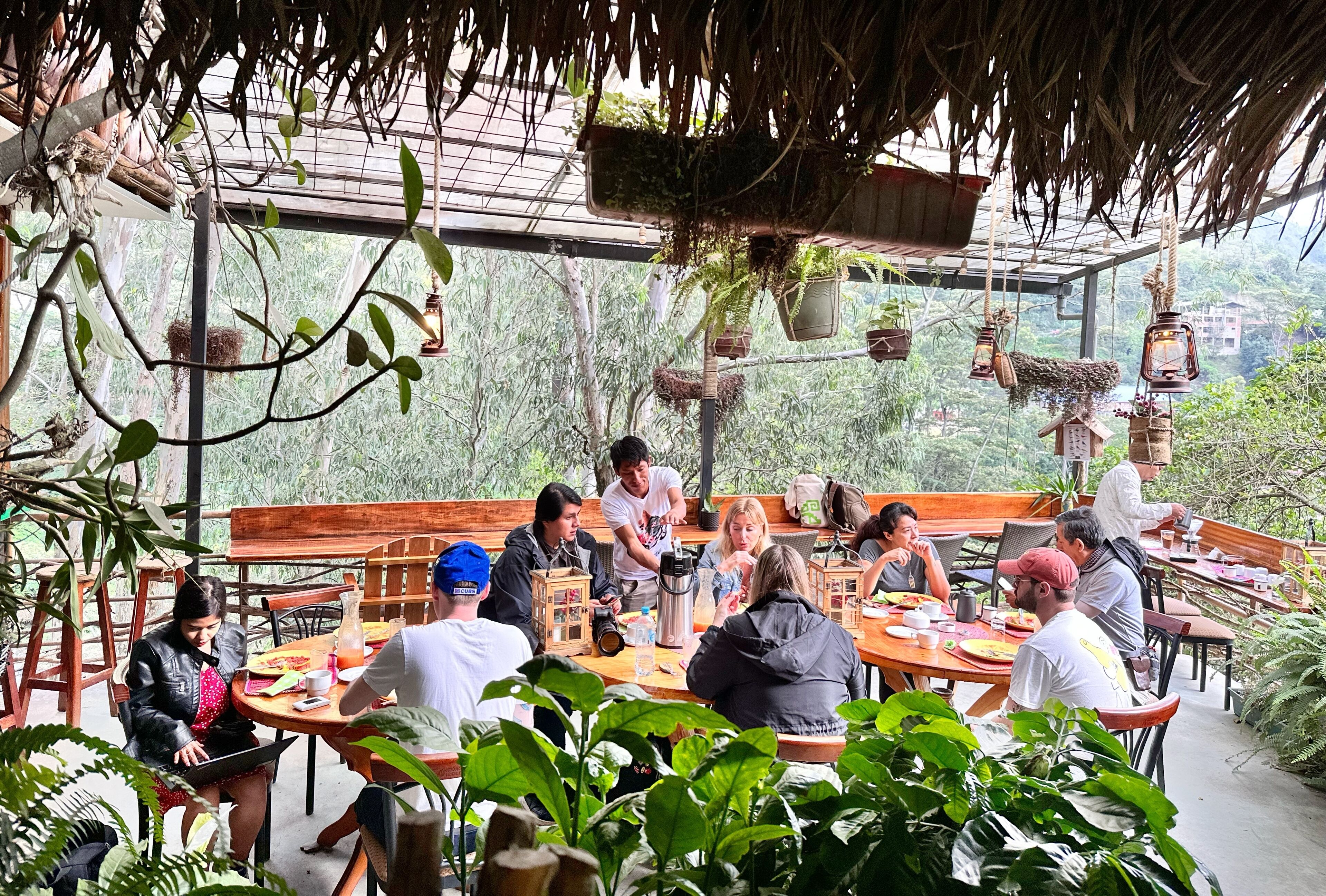 An outdoor dining area with wooden tables and chairs, surrounded by lush greenery and hanging planters.