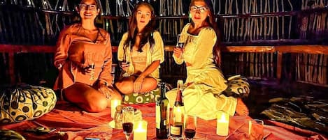 Three people seated on a bed with a table between them, holding drinks and surrounded by candles and bottles.