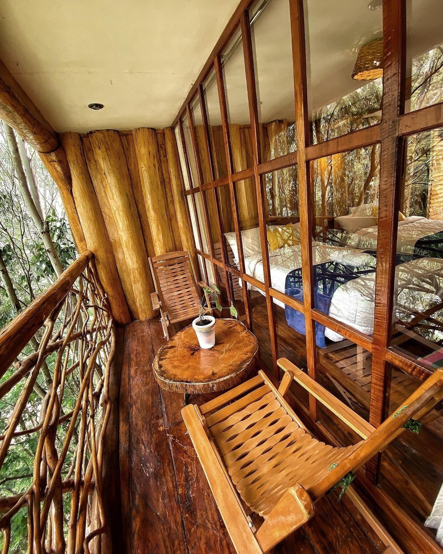 Une terrasse en bois avec une table et des chaises, un pot de fleurs et une fenêtre donnant sur les arbres.