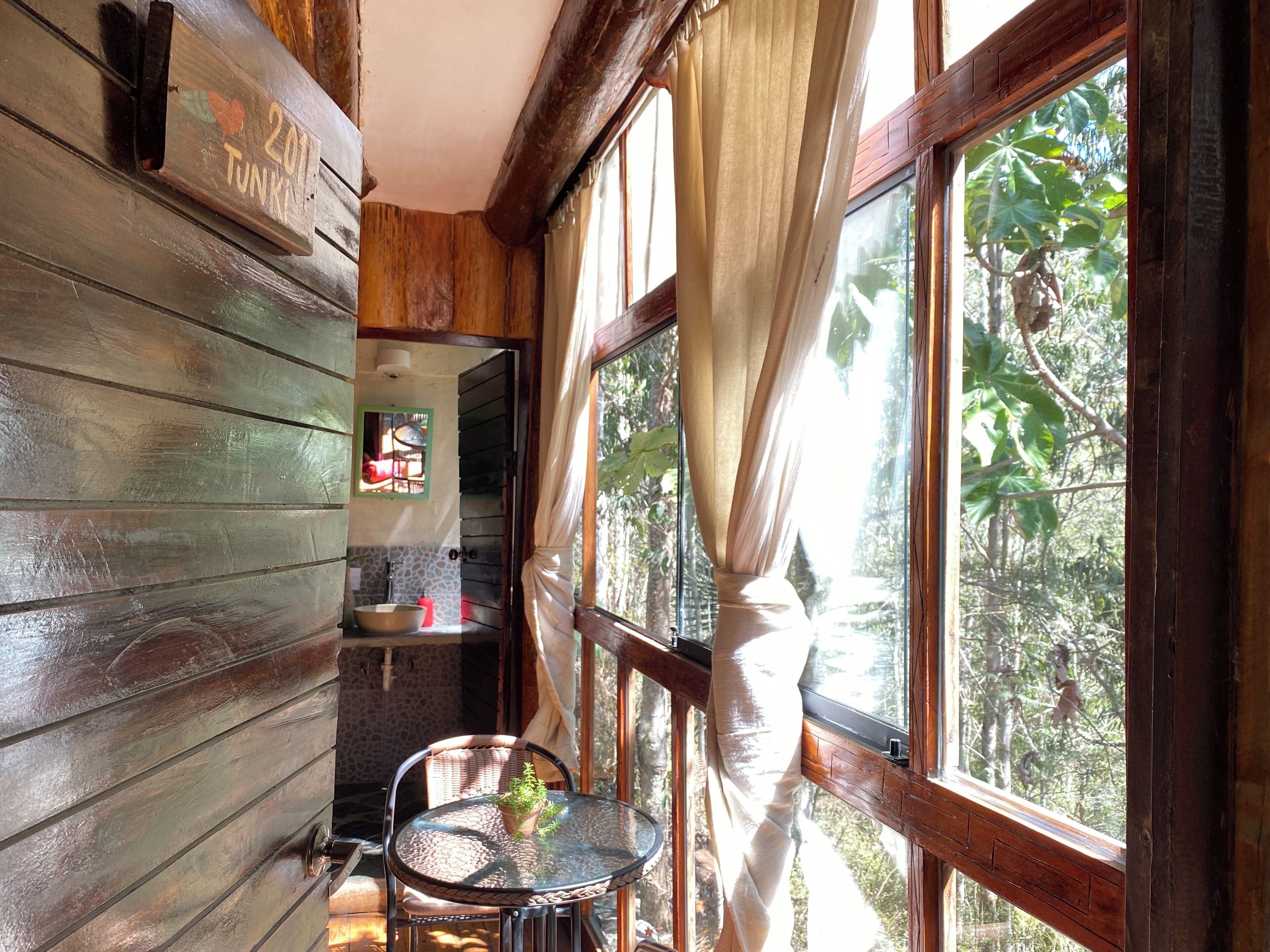 A wooden cabin interior with a dining area, a window with curtains, and a view of the outdoors.