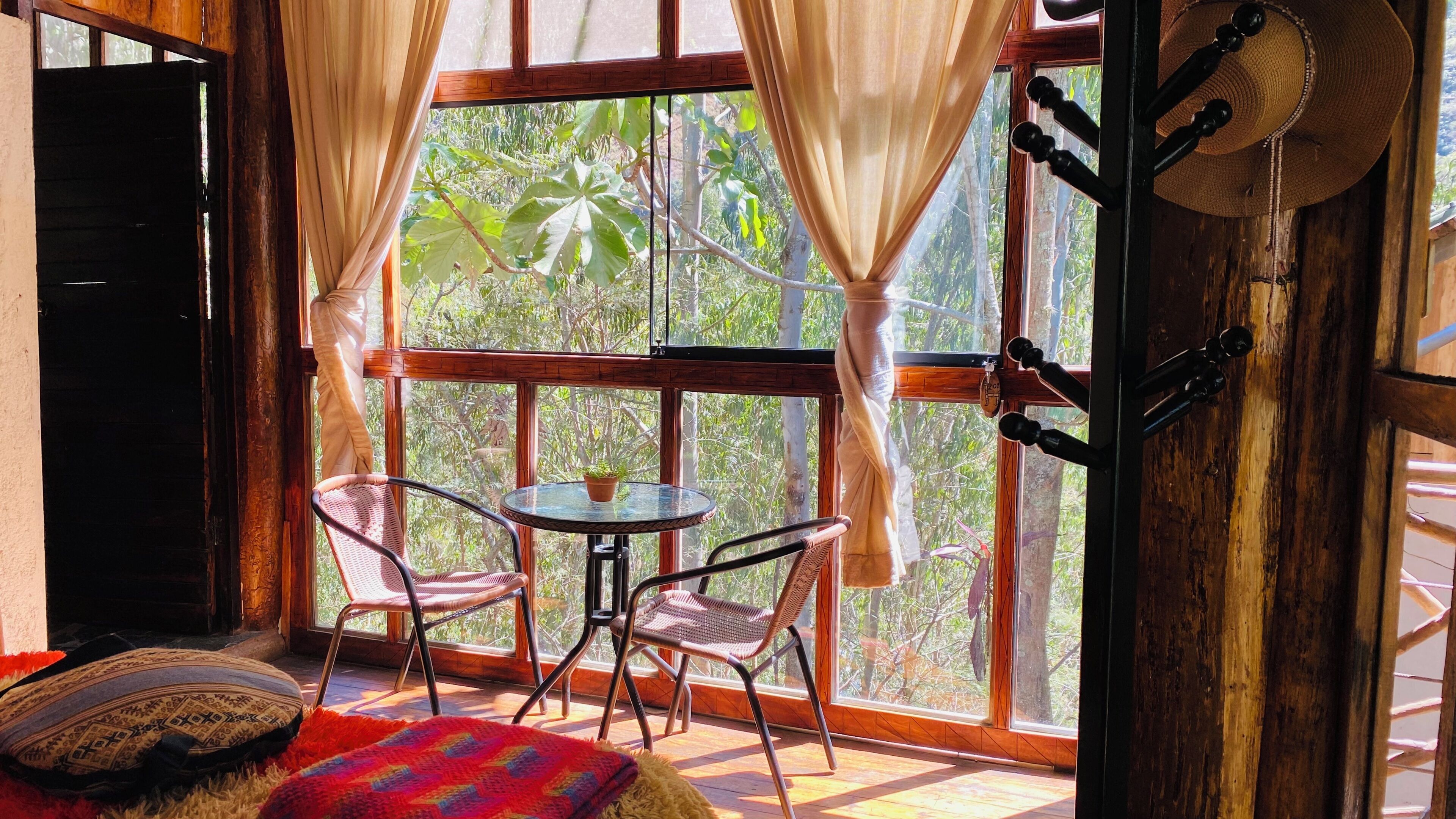 Una habitación con piso de madera, una mesa redonda con sillas, una ventana con cortinas y vistas a árboles.
