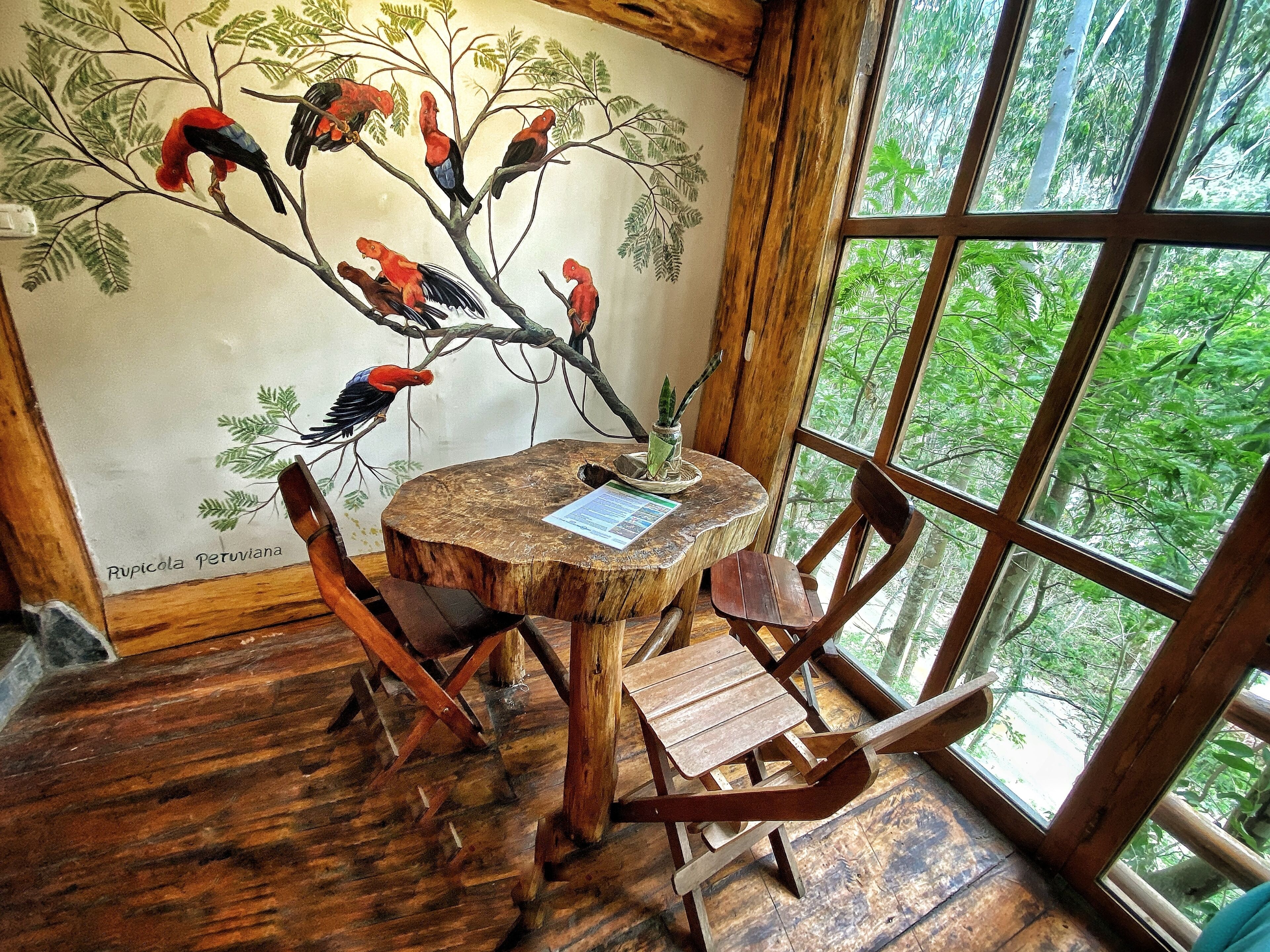 A rustic wooden interior with a round wooden table, chairs, and a wall mural of birds on a tree.