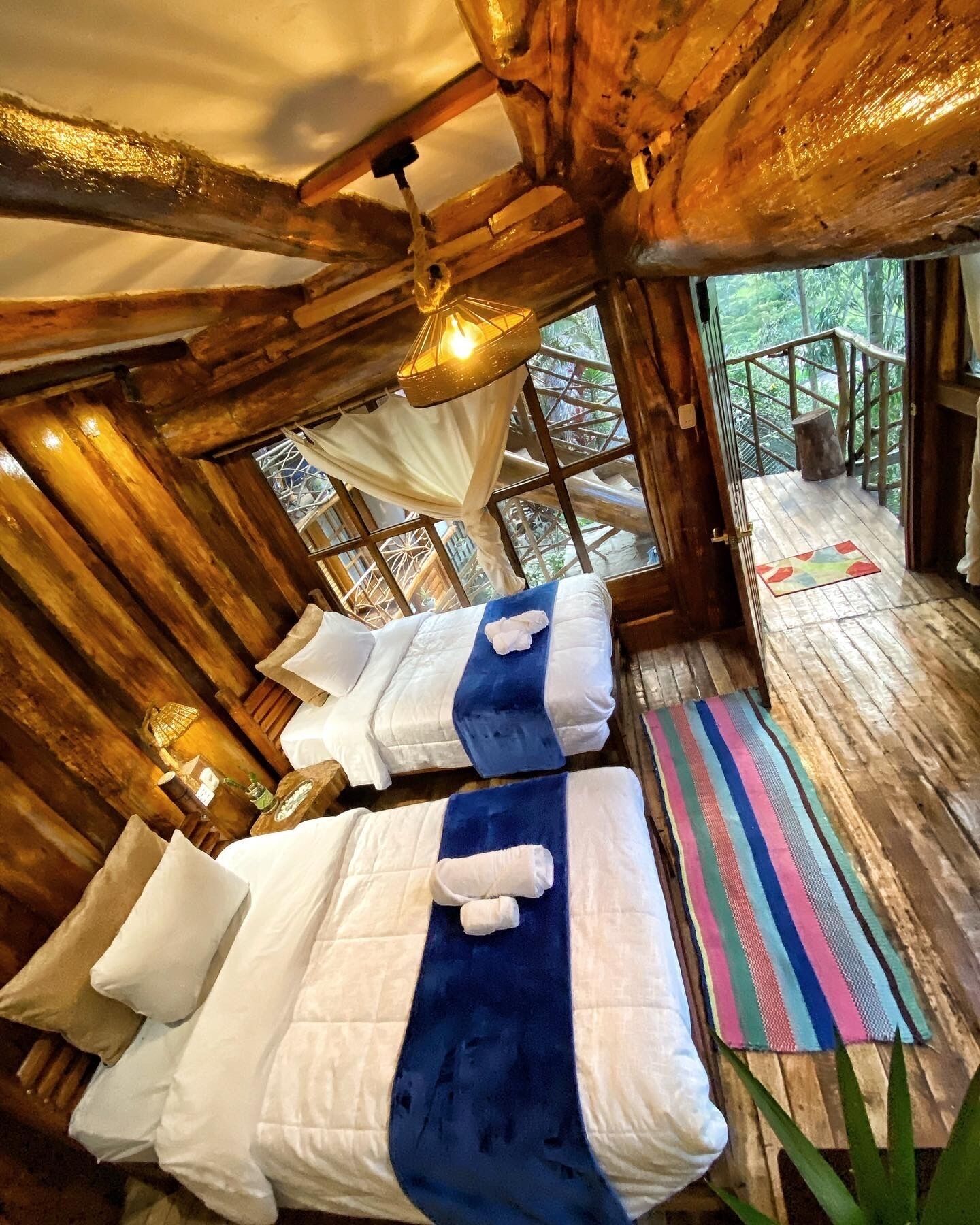 A wooden cabin with two beds, a hanging light fixture, and a view of the outdoors.