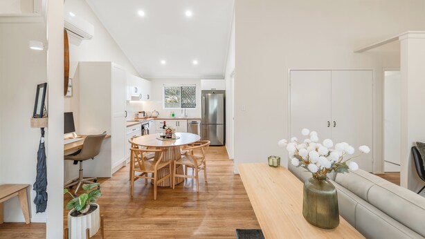 Una cocina moderna con armarios blancos, un comedor con una mesa redonda y sillas, y una sala de estar con un sofá y un jarrón de flores.
