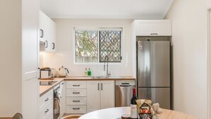 Cocina moderna con armarios blancos, refrigerador de acero inoxidable, ventana con vista a árboles y una mesa de comedor con vino y aperitivos.