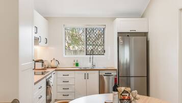 A modern kitchen with white cabinets, a stainless steel refrigerator, a window with a view of trees, and a dining table with a bottle of wine and snacks.