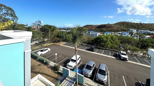 A parking lot with several cars, a building with a blue facade, and a view of a residential area with houses and hills in the background. - Trade Winds | Family Vacation PH | Near Ferry (Ceiba)