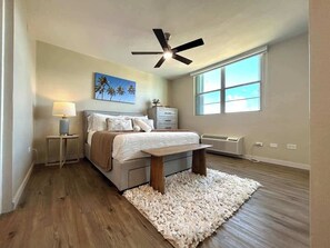 A bedroom with a bed, bedside table, lamp, ceiling fan, and a large window. - Trade Winds | Family Vacation PH | Near Ferry (Ceiba)