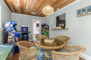 A room with a round glass-top table surrounded by wicker chairs, arcade games, and a wooden ceiling.
