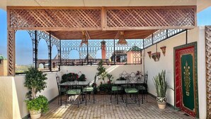 Un patio exterior techado con suelo de baldosas, mesas y sillas de metal, plantas en macetas y una puerta decorativa.