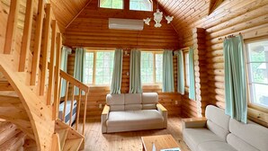 A log cabin interior with a wooden staircase, a sofa, a coffee table, and multiple windows with curtains.