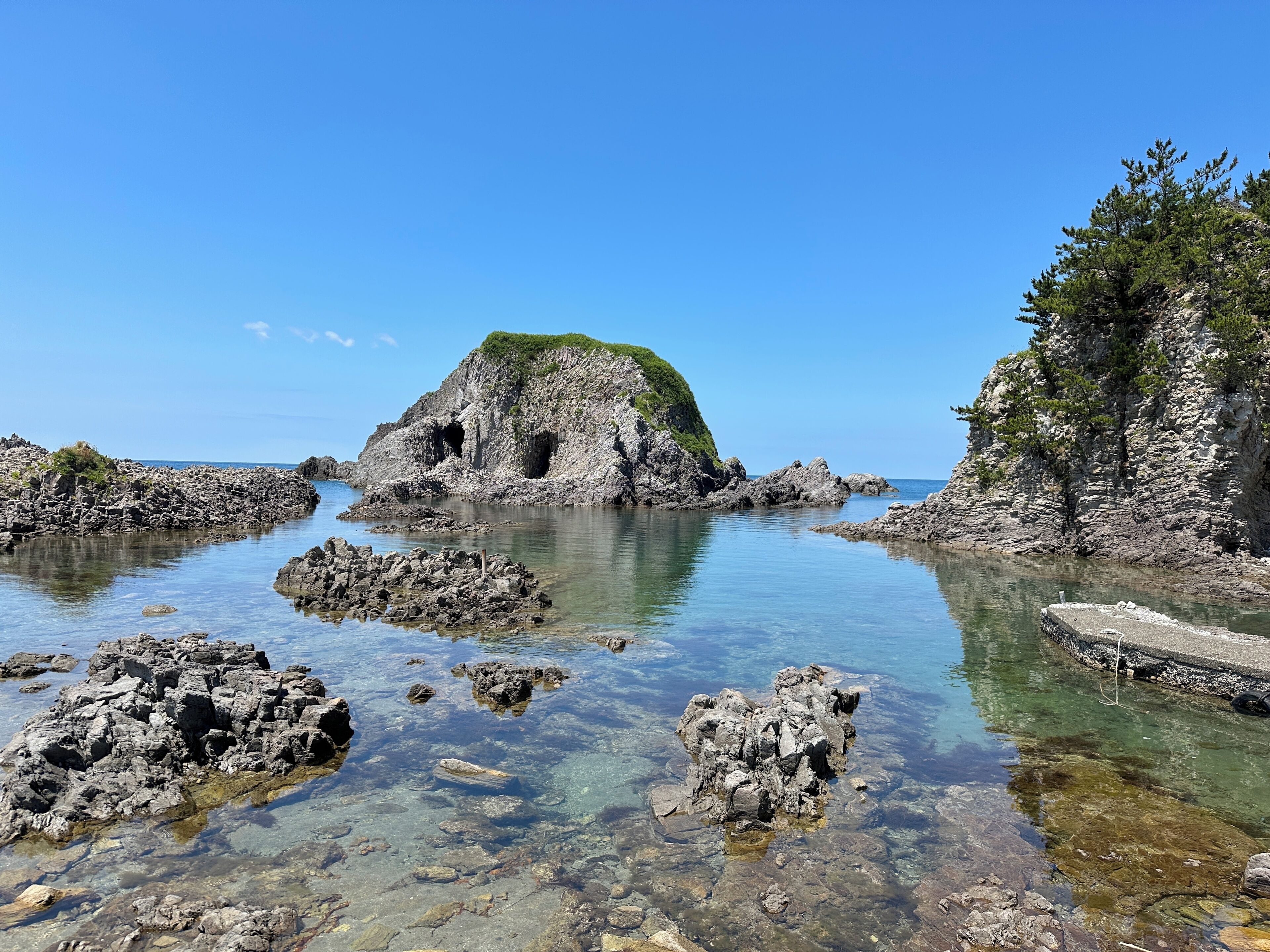 A rocky coastal landscape with clear water, a small cave-like structure, and a cliff with vegetation.