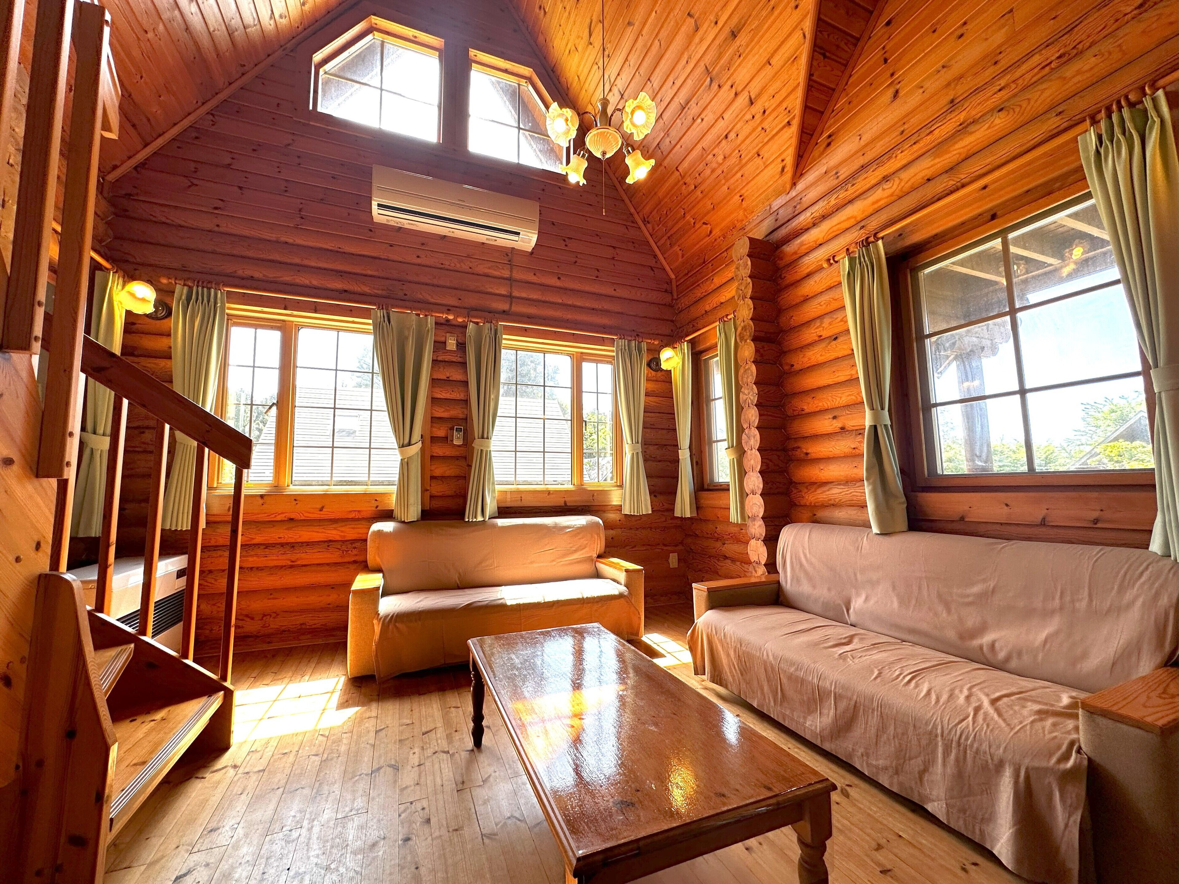 Interior de una cabaña de troncos con piso de madera, dos sofás, una mesa de madera y una escalera.