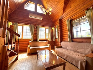 Interior de una cabaña de troncos con piso de madera, dos sofás, una mesa de madera y una escalera.