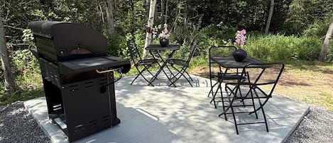 Zona de parrilla al aire libre con mesa y sillas, rodeada de árboles.