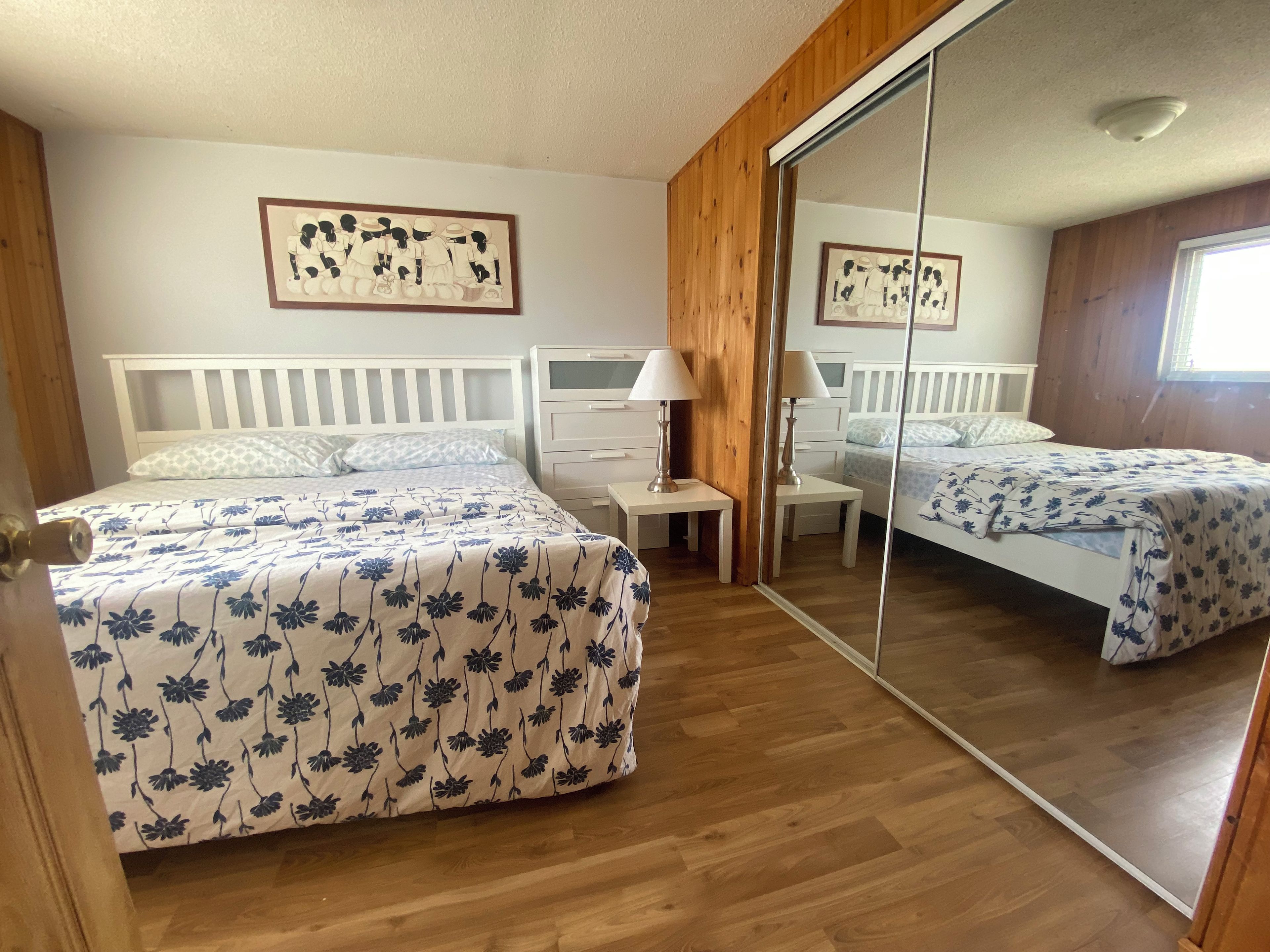A bedroom with two beds, a wooden wardrobe, a bedside table with a lamp, and a mirror.
