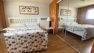 A bedroom with two beds, a wooden wardrobe, a bedside table with a lamp, and a mirror.