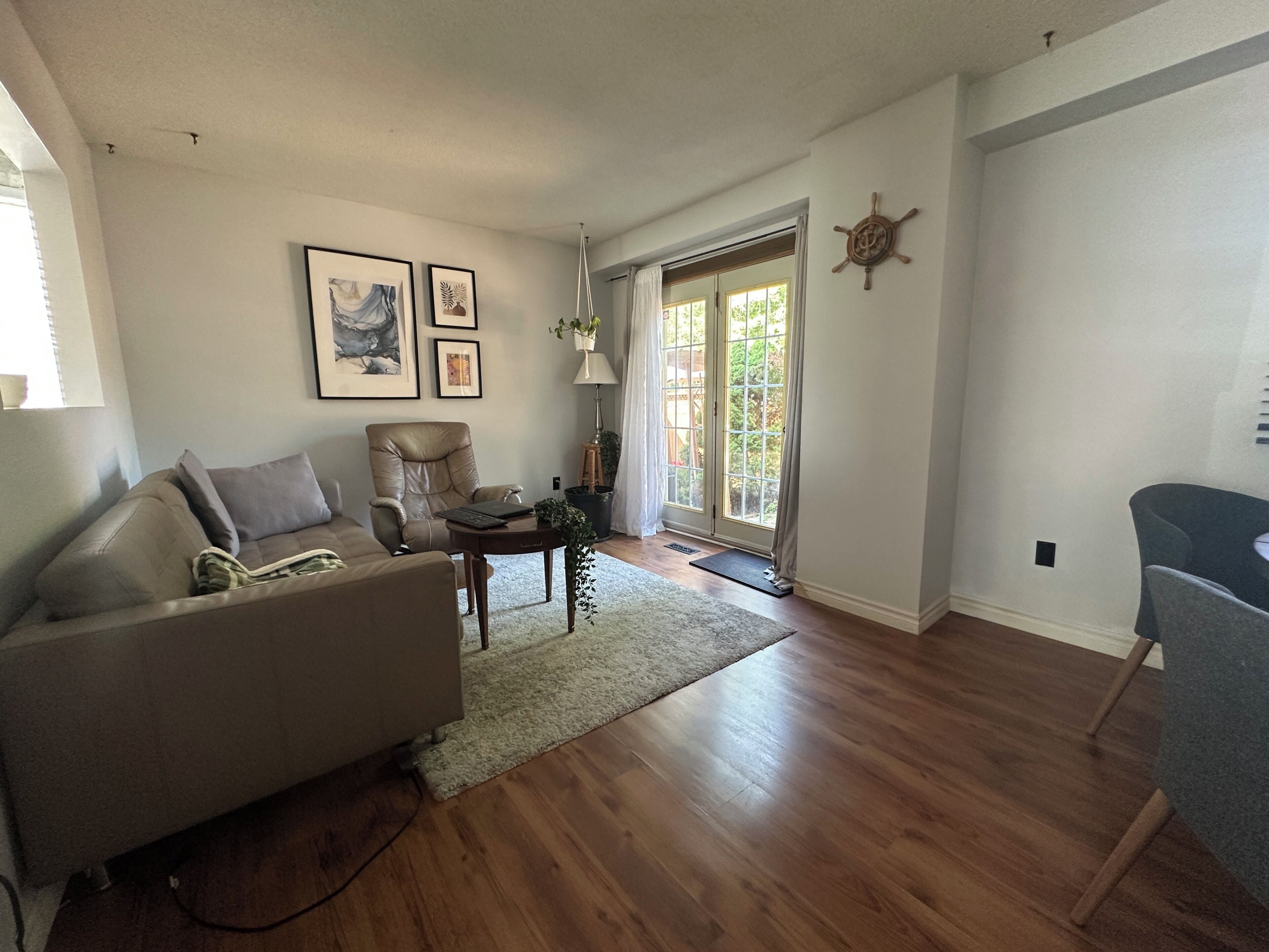 Una sala de estar con un sofá, un sillón, una mesita y una lámpara. En la pared hay cuadros enmarcados y una ventana con cortinas.