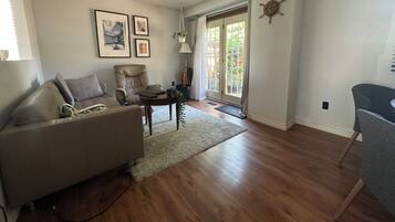 Una sala de estar con un sofá, un sillón, una mesita y una lámpara. En la pared hay cuadros enmarcados y una ventana con cortinas.