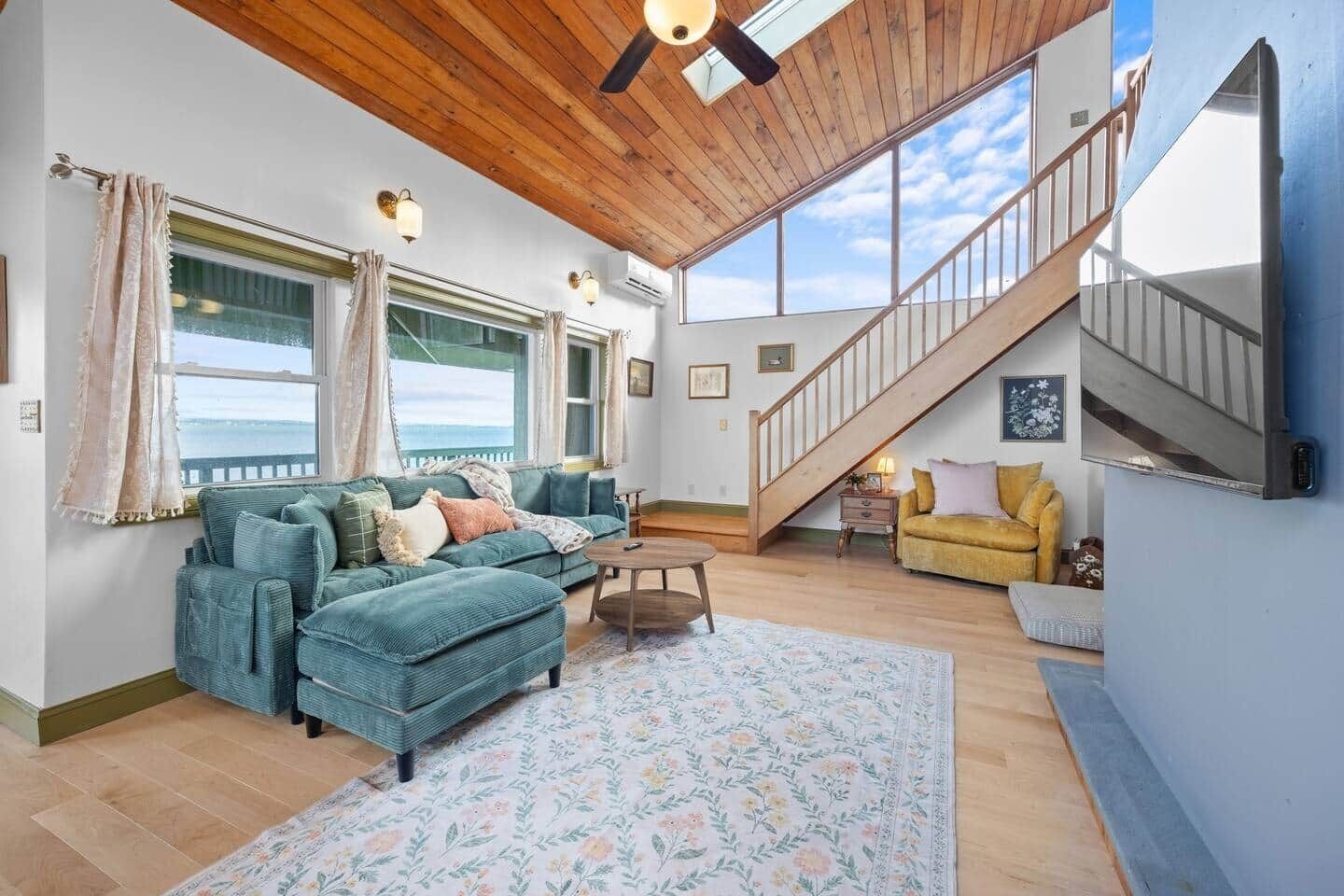 A spacious living area with a large window, a wooden ceiling, a staircase, and a sofa.