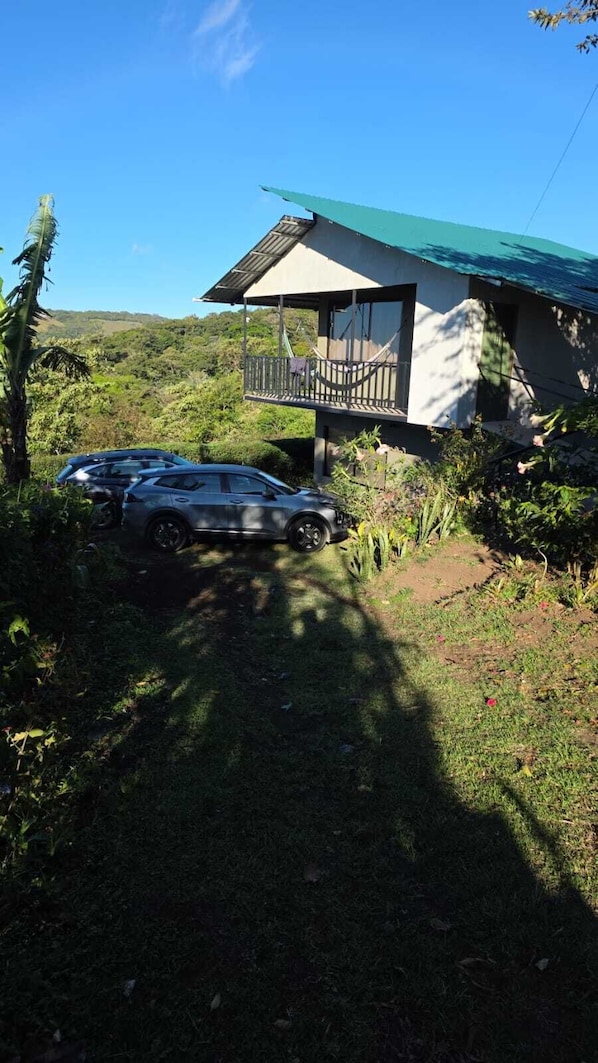 Una casa de dos pisos con un balcón, rodeada de vegetación y un coche estacionado.