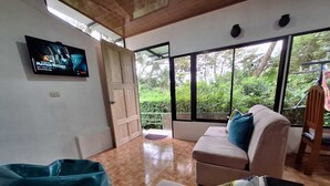 Een kamer met een televisie, een bank, een slaapzak en uitzicht op groen door grote ramen.