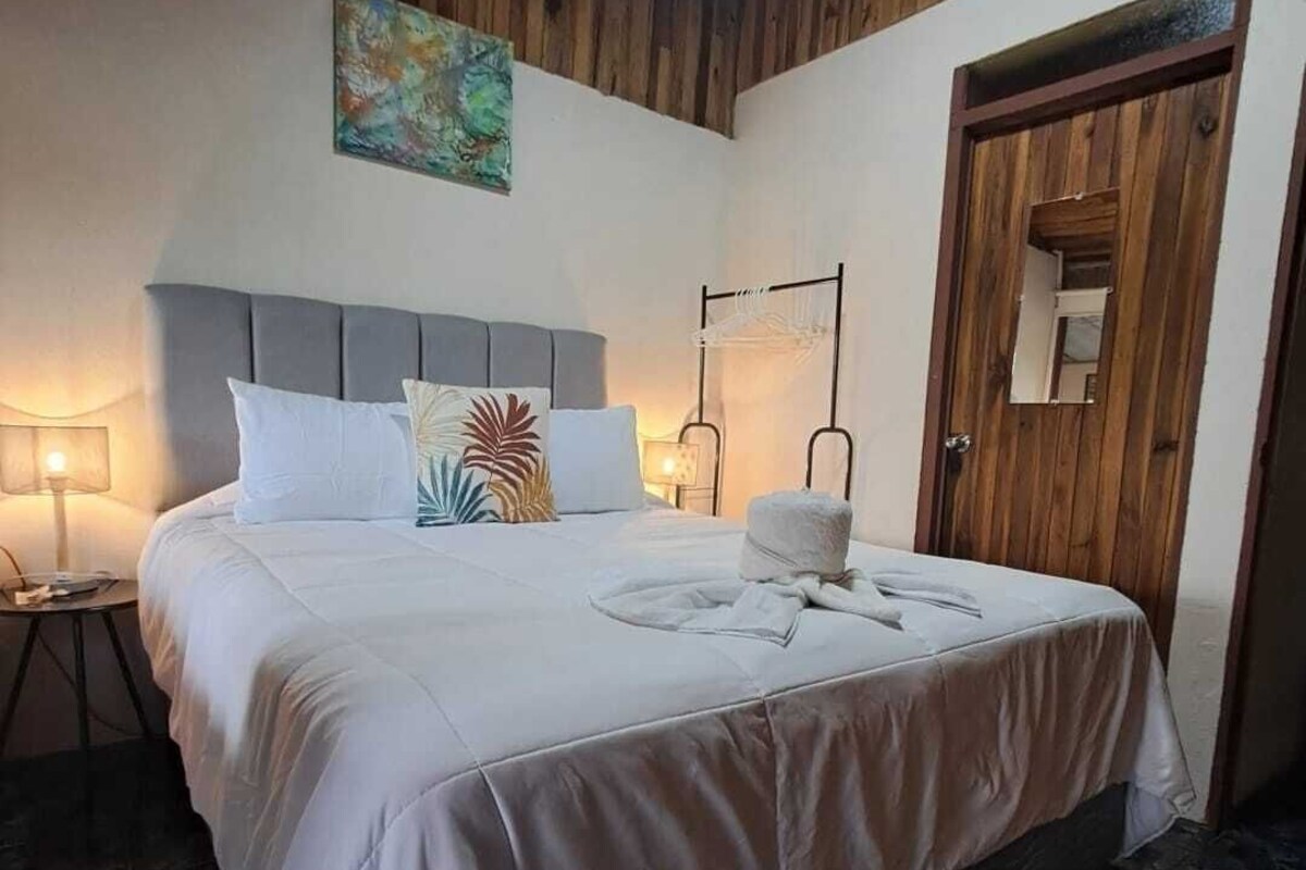 A bedroom with a bed, bedside lamps, a wooden ceiling, a door, and a painting on the wall.