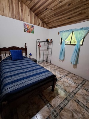 Un dormitorio con una cama, una ventana con cortinas, una estantería metálica y un cuadro en la pared.