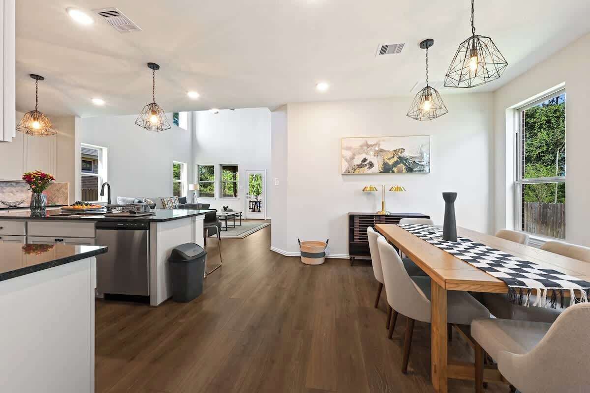 A modern kitchen with a central island, wooden flooring, pendant lighting, and a dining area with a checkered table runner.