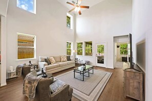 A spacious living room with a large sofa, a coffee table, and a television. There are multiple windows providing natural light and views of the outdoors. - Luxury 4BR 3BA - Peaceful Forest Retreat (Willis)