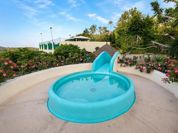 Ein türkisfarbenes Schwimmbecken mit Wasserrutsche, umgeben von üppiger Vegetation und rosa Blumen.