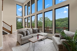 A spacious living room with a large window overlooking a forested area, a wooden staircase, and a cozy seating arrangement.