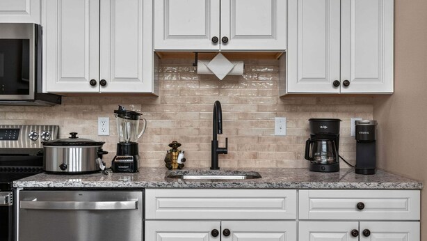 Una cocina moderna con armarios blancos, lavavajillas de acero inoxidable, cafetera y licuadora.