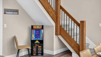 A wooden staircase with a handrail, a vintage Pac-Man arcade machine, and a single chair in a room with a light-colored wall and wooden flooring.