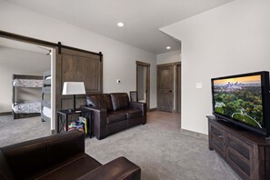 A living room with a brown leather sofa, a television on a wooden stand, and a cityscape view on the screen. - Aspen Trail Retreat - Breathtaking Mountain Views! (Estes Park)