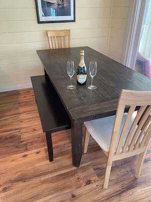 Une table de salle à manger en bois foncé, avec deux verres à vin et une bouteille de vin Korbel. Un banc en bois se trouve à gauche de la table, et une chaise avec un siège en osier est placée à proximité.