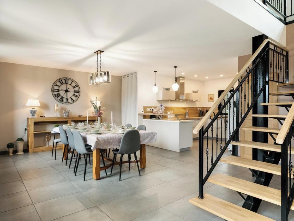 A modern kitchen with a dining area, a staircase, and a large wall clock.