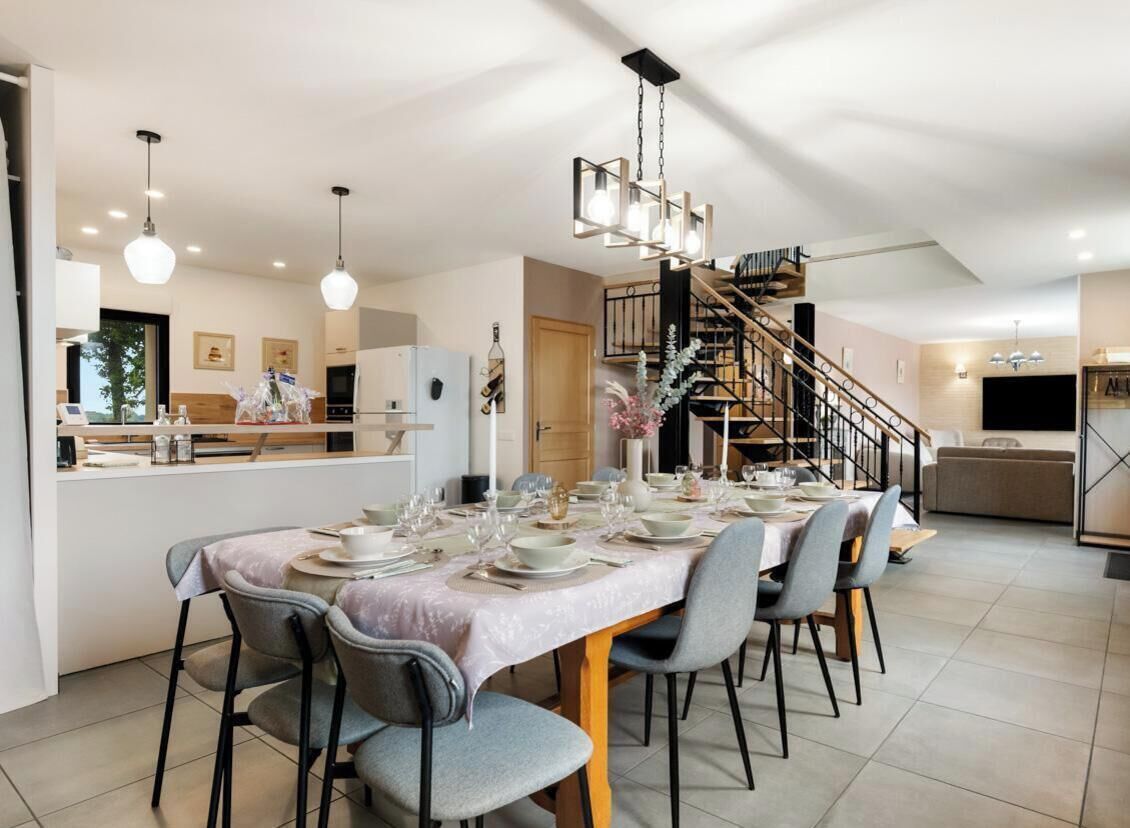A dining area with a long table set for six, a kitchen in the background, and a staircase leading to an upper level.