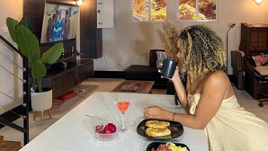 En person sitter vid ett bord med mat och en dryck i ett rum med träbjälkar, en tv och konstverk på väggen.