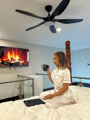 A person sitting on a bed with a tablet, holding a mug, in a room with a ceiling fan, a TV displaying a fireplace, and a railing.