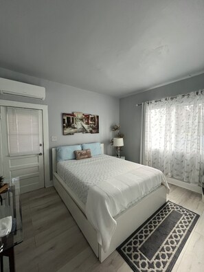 A bedroom with a bed, a bedside table, a lamp, a window with curtains, and a door.